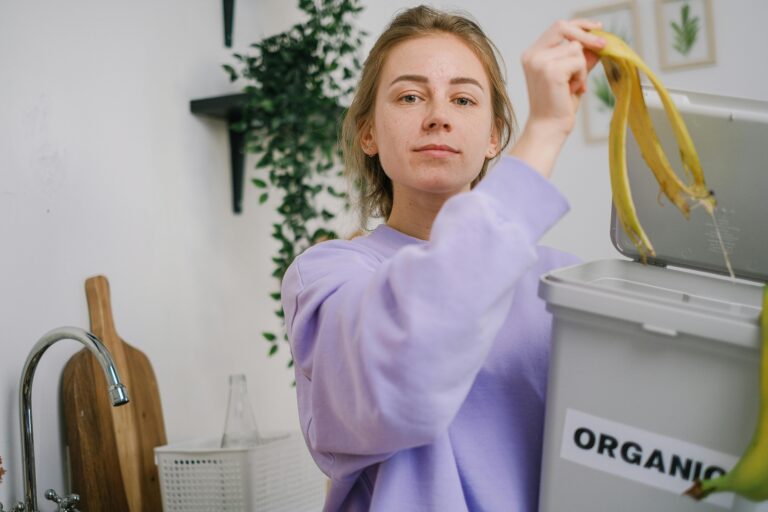 “A woman in a purple sweatshirt placing banana peels into a kitchen bin labeled ‘organic,’ demonstrating a simple DIY method for making beginner-safe liquid fertilizer.
