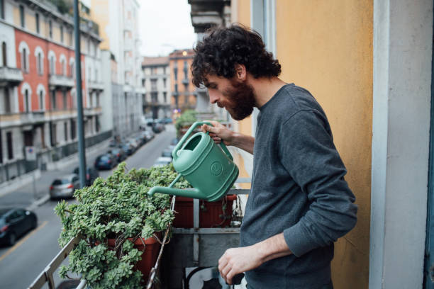 Man watering balcony plants using a green watering can, showing proper container selection and balanced watering routine for small urban gardens.