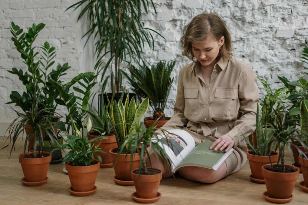 A woman sitting on the floor reading a gardening book surrounded by indoor potted air-purifying plants such as snake plant, peace lily, and ZZ plant, showing real small-space balcony gardening care and plant setup ideas.